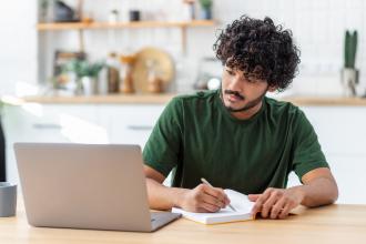 Man writing notes while taking an online class on computer.