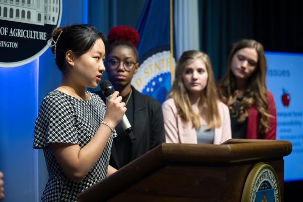 A female college student standing behind a podium and speaking into a microphone, with three other female students looking on.